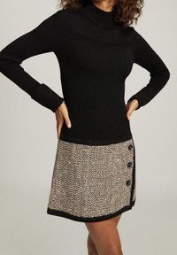 Pull à col roulé côtelé noir associé à une jupe beige texturée avec un ourlet noir et trois boutons décoratifs sur le côté.