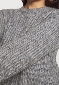 Szary sweter z dzianiny o ribbowej fakturze i okrągłym dekolcie, wykonany z wełny średniej grubości. Zbliżenie pokazuje detale rękawów.