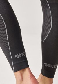 Black athletic leggings with textured fabric, featuring gray accents and the brand name "SNOCKS" printed on the lower leg.