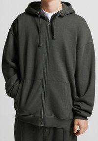 Sudadera con cremallera de color gris oscuro hecha de un suave tejido de algodón. Presenta una capucha, bolsillos frontales y puños acanalados en las mangas y el dobladillo.