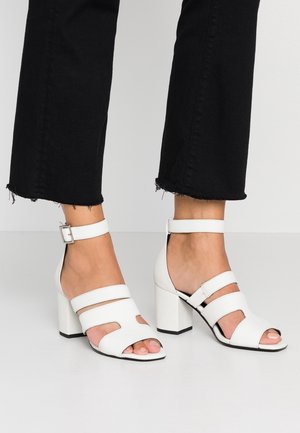 Sandales blanches à talons blocs ouvertes à l'avant avec brides à la cheville, portées avec un pantalon noir court à ourlet effiloché sur un sol gris clair.