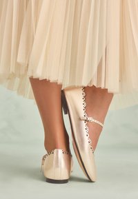 Chaussures plates métalliques dorées avec un bout rond, un bord festonné et une petite bride à boucle, associées à une jupe en tulle nude à plusieurs couches.