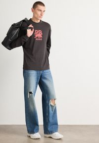 Maglietta nera a maniche lunghe con logo rosso, jeans azzurri chiari strappati e sneakers bianche. Il modello porta una borsa nera.