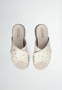 Sandali scivolati beige con fascia incrociata e motivi geometrici dorati, suola piatta e suola testurizzata per una buona aderenza. Materiale superiore synthético liscio.