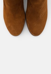 Brązowe zamszowe botki za kostkę z zaokrąglonym noskiem i gładką teksturą, charakteryzujące się minimalistycznym stylem i brakiem widocznych elementów metalowych oraz ozdób.