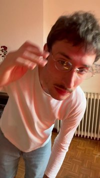 Fiatal férfi szemüvegben, fehér hosszú ujjú ingben és farmerben, előrehajolva, kezét felemelve egy belső térben, egy radiátor és növény közelében.