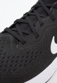 Černá sportovní bota s texturovaným textilním svrškem, s černými šněrovadly a bílým logo. Design zahrnuje perforované detaily a bílou podešev.