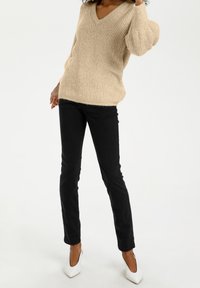 Pull beige en tricot avec encolure en V, manches oversize, associé à un jean noir ajusté et des chaussures à talons blanches. Texture lisse.