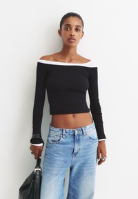 Schwarzes langärmliges Crop-Top mit weißen schulterfreien Akzenten, kombiniert mit hellblauen weiten Jeans. Glatte Textur, taillierte Passform, lässiger Stil.