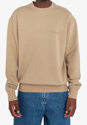 Mann trägt ein beiges Langarm-Sweatshirt mit Rundhalsausschnitt und "Element"-Logo auf der Brust sowie blaue Jeans, steht vor einem einfarbigen Hintergrund.