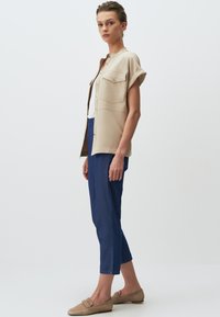 Beżowa koszula krótko rękawowa w stylu kurtki z dwoma kieszeniami na piersi, z przodem na guziki, połączona z niebieskimi spodniami o prostym kroju i beżowymi butami wsuwanymi.