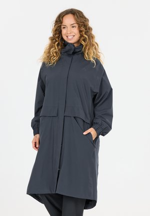 Kvinde med krøllet hår iført en lang, mørkegrå hættetrøje med elastiske manchetter og hænder i forlommerne, smilende og kiggede til siden.