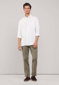 Camisa de lino blanca con botones y mangas enrolladas, combinada con pantalones verdes oliva y zapatos de ante marrón, de pie sobre un fondo neutro.