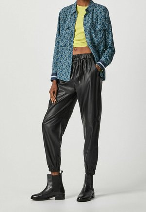 Person trägt blau gemustertes Hemd über gelbem bauchfreiem Top, schwarze Jogger-Hosen aus Leder und schwarze Stiefeletten, steht vor weißem Hintergrund.