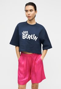 Μπλε ναυτικό κοντομάνικο μπλουζάκι με λευκό κείμενο "LALA BERLIN"· συνδυασμένο με ροζ σατέν σορτς με ελαστική μέση και τσέπες.