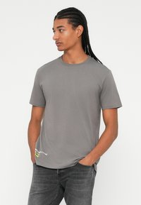 Camiseta gris de manga corta hecha de algodón. Cuello redondo con un pequeño detalle gráfico en la parte inferior izquierda. Ajuste relajado.
