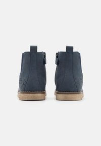 Ett par marinblå ankestävlar med dragflikar, bakstygn och lätta trästruktur-sulor, visade från baksidan på vit bakgrund.