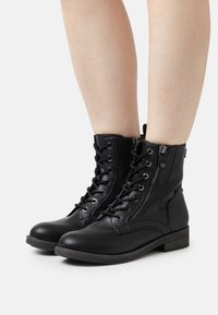 Botas negras hasta el tobillo de cuero texturizado con puntera redonda, cremallera vertical y cordones en la parte delantera. Cuenta con un tacón bajo y cuadrado.