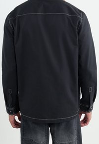 Černá košile s dlouhými rukávy a kontrastním bílým prošíváním, volný střih a knoflíkové manžety. Má jednoduchý design na zadní straně.