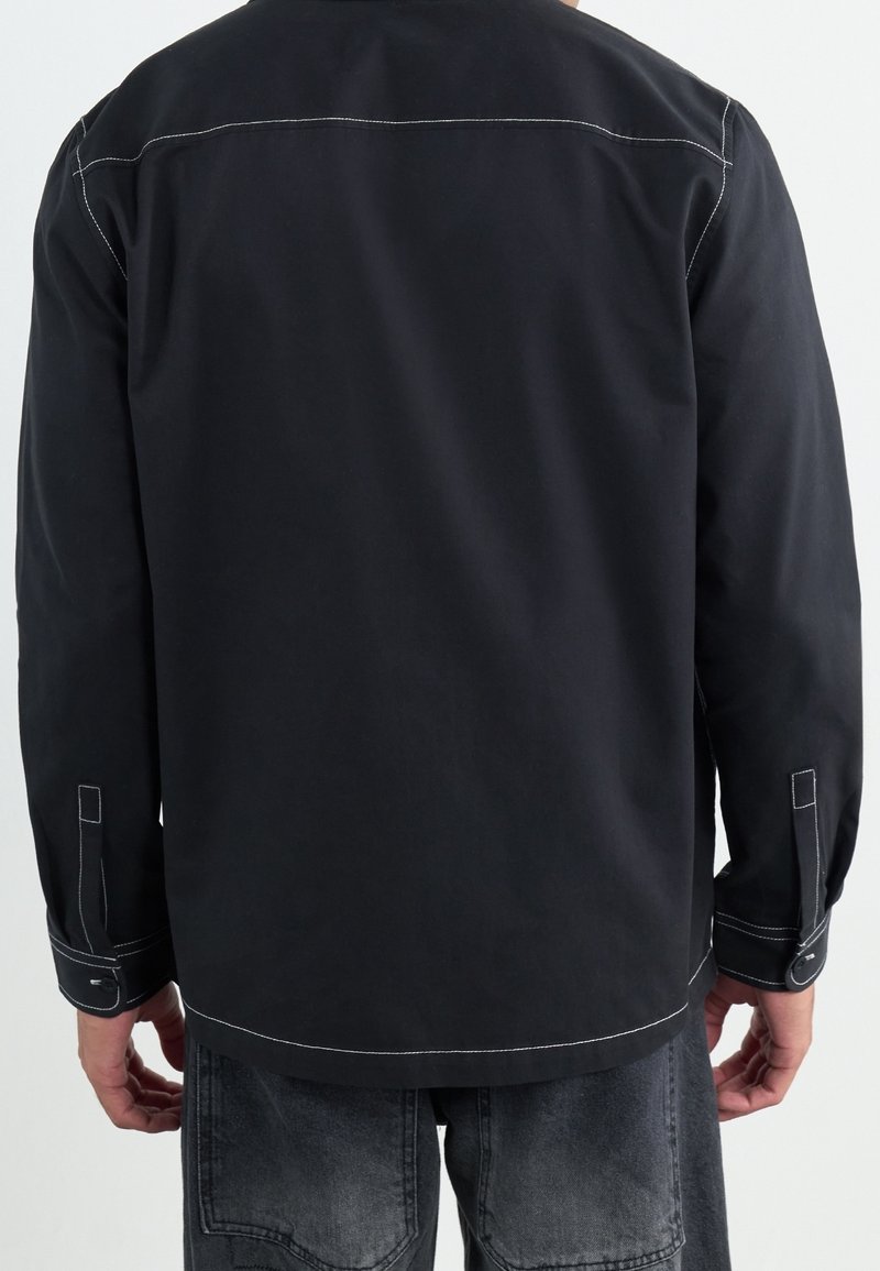 Černá košile s dlouhými rukávy a kontrastním bílým prošíváním, volný střih a knoflíkové manžety. Má jednoduchý design na zadní straně.