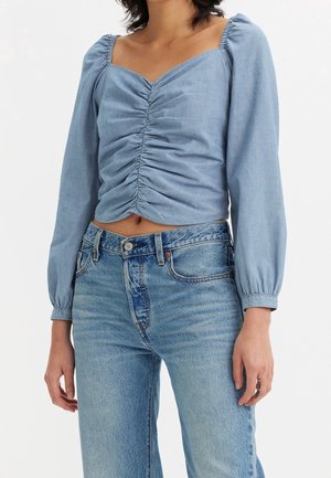 Femme portant un haut bleu clair froncé à manches longues avec un col carré et un jean bleu délavé taille haute sur fond blanc.
