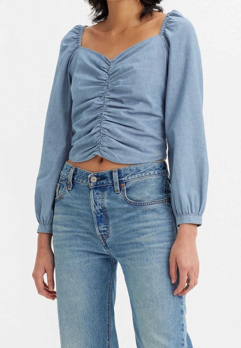 Femme portant un haut bleu clair froncé à manches longues avec un col carré et un jean bleu délavé taille haute sur fond blanc.