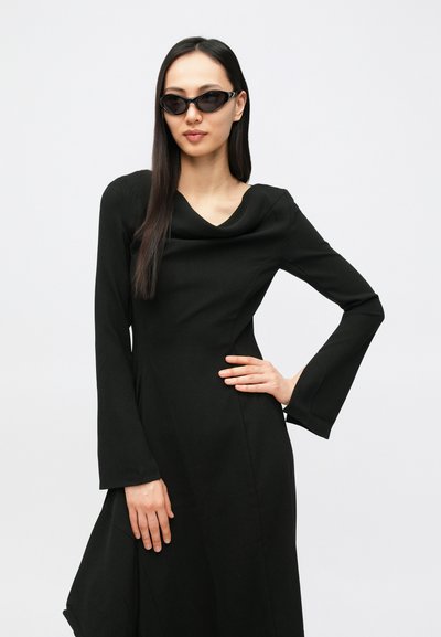 Μαύρο φόρεμα με μακριά μανίκια, ντεκολτέ με σκίσιμο και φουσκωτά μανίκια. Το μοντέλο φορά κομψά μαύρα γυαλιά ηλίου, τα μαλλιά είναι ίσια και ανοιχτά.