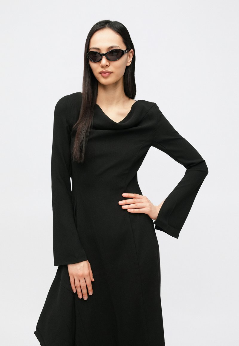 Vestito nero a maniche lunghe con scollo drappeggiato e maniche svasate. La modella indossa occhiali da sole neri eleganti, capelli lisci e sciolti.