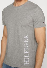Graues Baumwoll-T-Shirt mit kurzen Ärmeln, verfügt über vertikalen weißen "HILFIGER"-Text an der Seite und ein kleines Logo-Detail am Ärmel.