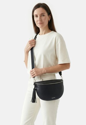 Woman in white outfit holding a black leather crossbody bag with zipper and tassel detail on a plain background.