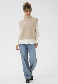 Beige knit sweater vest with a turtleneck, featuring a textured diamond pattern, layered over a white blouse and paired with light blue jeans.