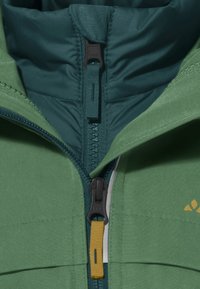 Veste verte avec un col haut, dotée d'une fermeture éclair proéminente avec une tirette jaune et un tissu texturé. Petit logo dans le coin supérieur.