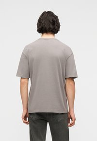 Άνδρας με σκούρα μαλλιά, φορώντας μια απλή γκρι κοντομάνικη μπλούζα και σκούρα γκρι τζιν, με την πλάτη προς τα εμπρός, μπροστά σε απλό φόντο.