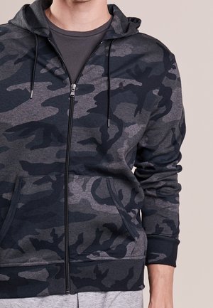 Mężczyzna noszący ciemnoszarą i czarną bluzę z kapturem na suwak z troczkami i przednimi kieszeniami, pod którą ma jednolitą ciemnoszarą koszulkę.