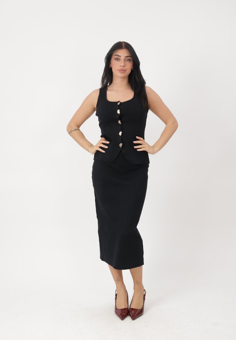 Vrouw met lang donker haar, gekleed in een zwarte mouwloze top met knopen en een bijpassende lange rok, poserend met handen in de zij en rode hakken.