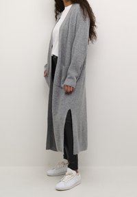 Gray ribbed cardigan with long sleeves, open front, and side slits. Worn over a white top and paired with black pants and white sneakers.