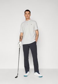 Lys grå polo skjorte med tre knapper, mørk grå bukser, hvite golf sko med blå detaljer, holder en golfklubbe, har på seg en hvit hanske.