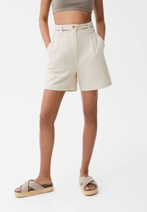 Kvinde iført beige højtaljede shorts med nålestriber og sidelommer samt beige platformsandaler med krydsremme, står med hænderne i lommerne.