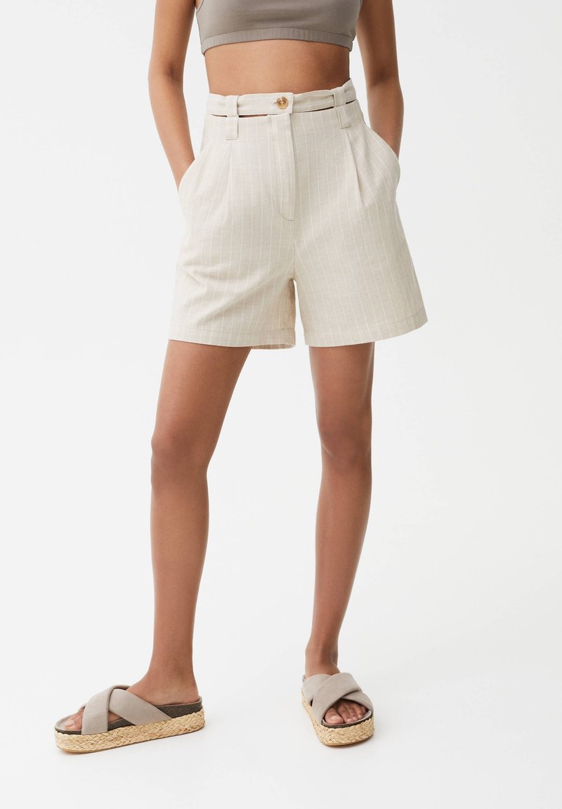 Femme portant un short beige à rayures fines taille haute avec poches latérales et des sandales compensées beiges à brides croisées, debout les mains dans les poches.