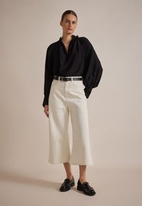 Zwarte blouse met lange mouwen en geplooide schouders, gecombineerd met crèmekleurige, wijde broek en zwarte instappers. Eenvoudige riem met zilveren gesp.