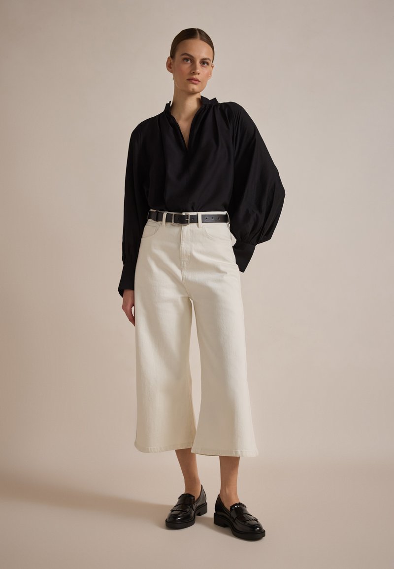 Zwarte blouse met lange mouwen en geplooide schouders, gecombineerd met crèmekleurige, wijde broek en zwarte instappers. Eenvoudige riem met zilveren gesp.