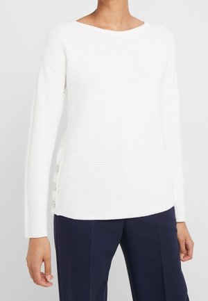 Hvid langærmet ribstrikket sweater med bådudskæring og sølvknapper i siden, båret med marineblå skræddersyede bukser.