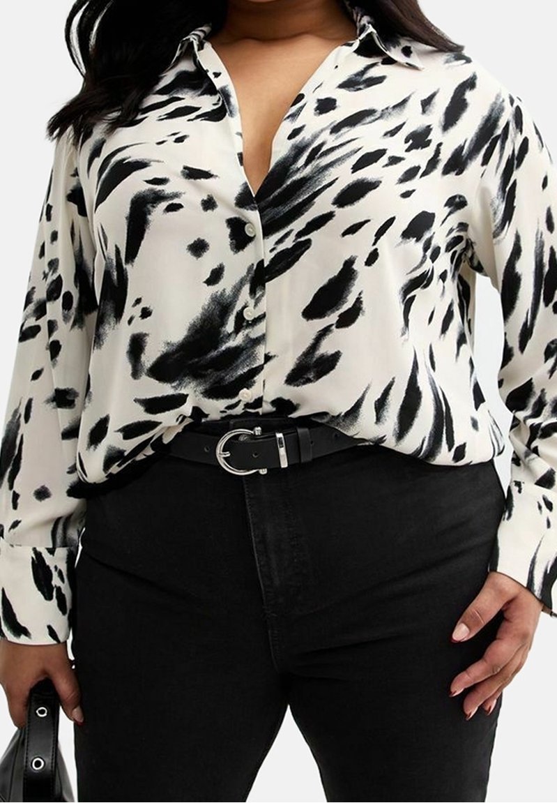 Witte blouse met zwart abstract patroon, lange mouwen, knoopsluiting aan de voorkant en kraag. Gepaard met zwarte jeans en een zwarte riem.