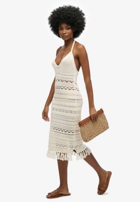 Häkelt Maxikleid in Off-White mit V-Ausschnitt, gemustertem Material und Fransen-Saum. Kombiniert mit einer geflochtenen Clutch und braunen Sandalen.