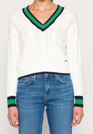 Pull blanc en maille torsadée à col en V avec des rayures vertes et noires au niveau de l'encolure et des poignets, associé à un jean en denim bleu.