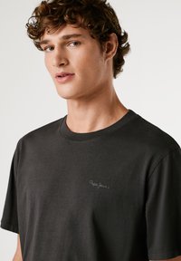 Tricou scurt de culoare gri închis, confecționat din bumbac moale. Are un guler rotund și un mic logo brodat pe piept. Textură netedă.