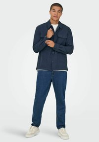 Camisa azul marino abotonada con dos bolsillos en el pecho, combinada con jeans azules y zapatillas blancas. La camisa tiene cuello y mangas largas.