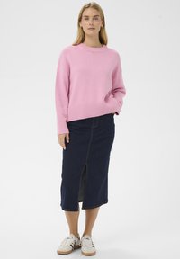 Pull rose tricoté avec ourlet et poignets côtelés, associé à une jupe en jean foncé avec une fente à l'avant. Des baskets blanches complètent la tenue.