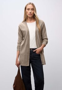Cardigan largo de color beige claro con bordes acanalados, de frente abierto, combinado con una blusa blanca y jeans de color azul oscuro, llevando un bolso marrón.