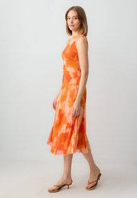 Orange Batik-Top ohne Ärmel, kombiniert mit einem fließenden, plissierten Rock. Der Stoff ist leicht, hat eine weiche Textur und ein lebhaftes, gemischtes Muster.
